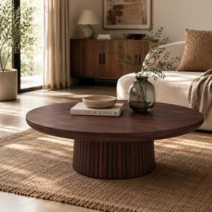 A round dark wood coffee table with a fluted pedestal base, styled in a bright, modern living room with a woven rug, a beige sofa, and a dark glass vase.
