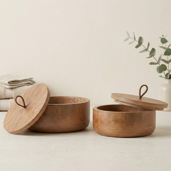 Two wooden storage bowls with lids on a light surface with a plant in the background.