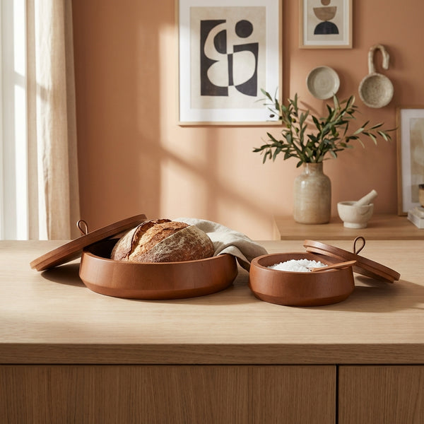 Wooden bread box with a loaf of bread on a wooden table, with a vase and decorative items in the background.