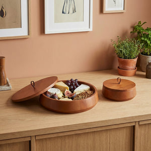 Wooden food platter with snacks on a wooden surface with plants and framed pictures in the background.