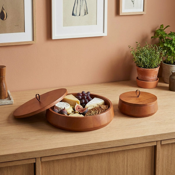 Wooden food platter with snacks on a wooden surface with plants and framed pictures in the background.