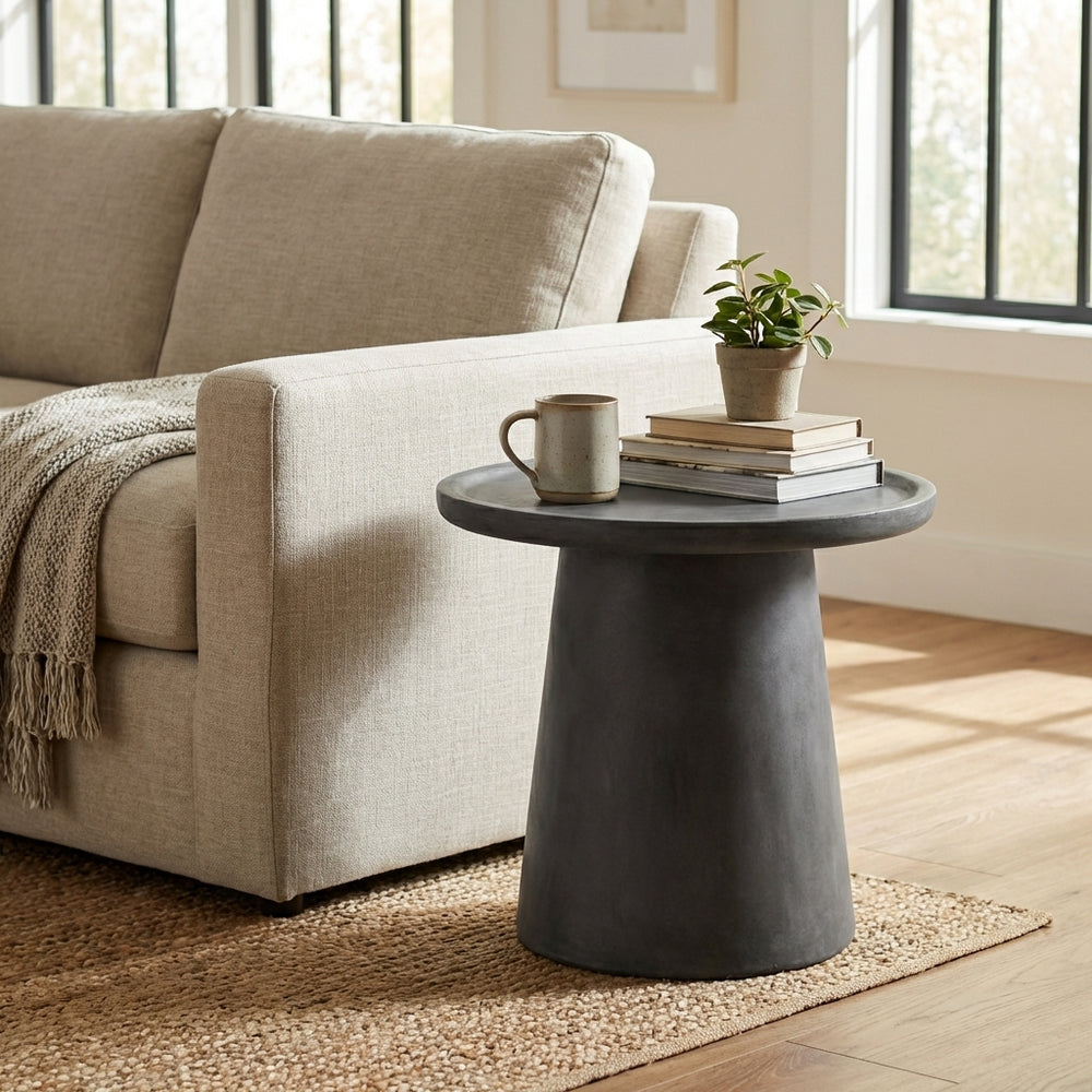 Beige sofa with a small round dark gray table holding books and a mug, in a bright room.