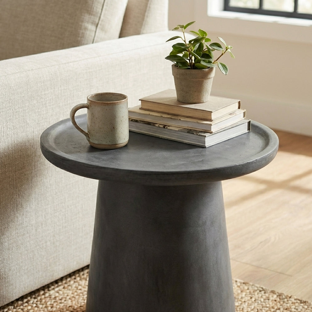 Beige sofa with a small round dark gray table holding books and a mug, in a bright room.