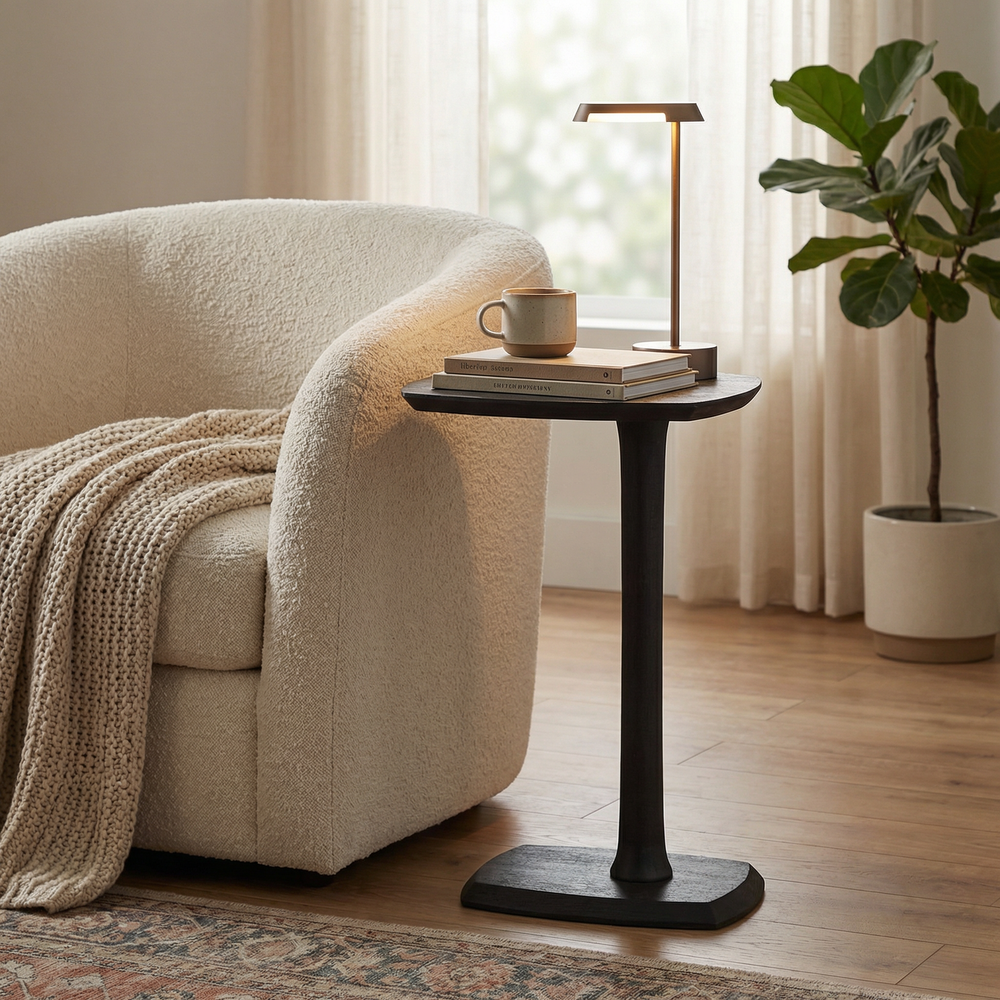 Small black side table with a cup, books, and plant next to a beige armchair in a living room.