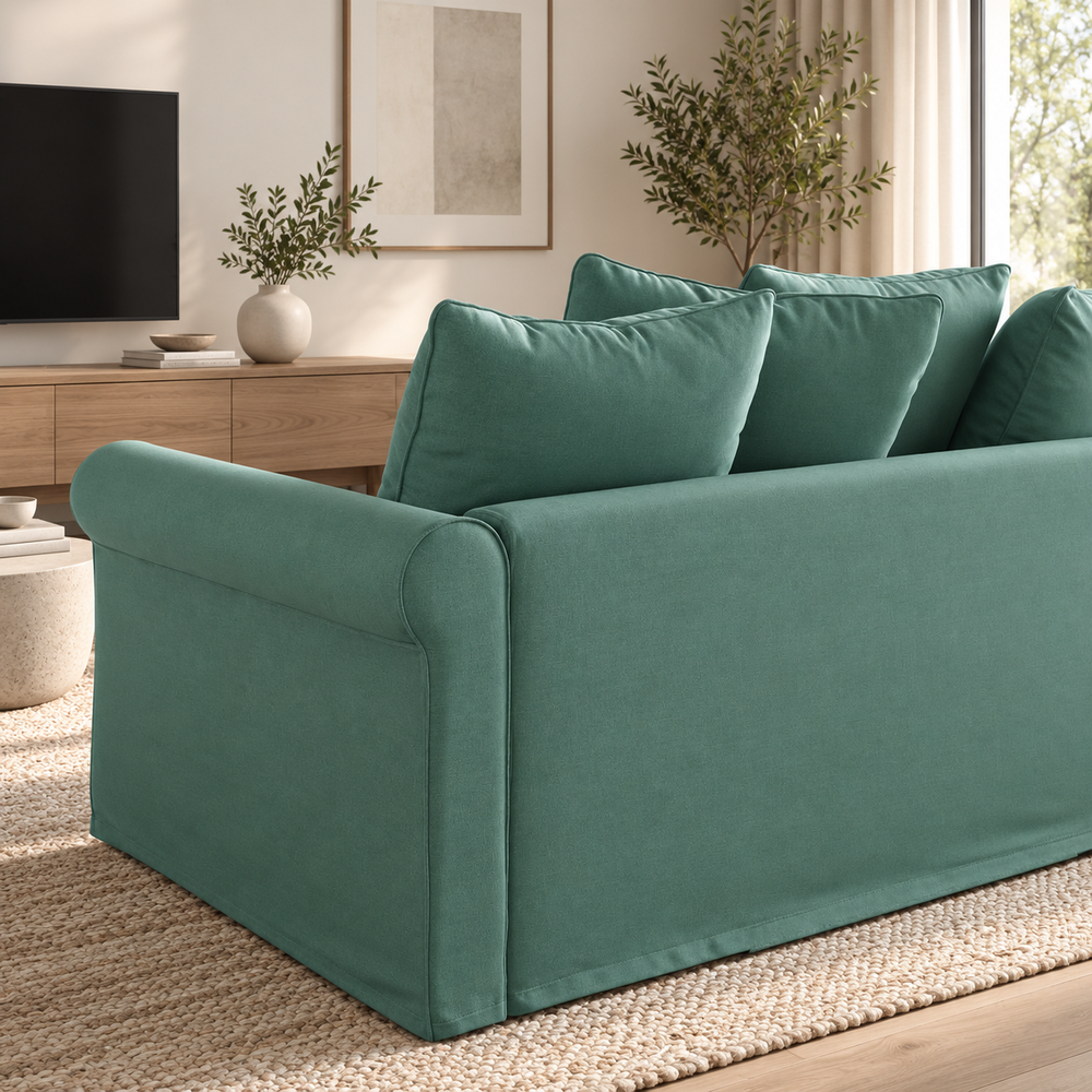 A green sectional sofa with a chaise lounge in a bright, modern living room. The sofa features plush cushions, a matching throw blanket draped over one arm, and is styled on a textured white rug with a light wood floor.