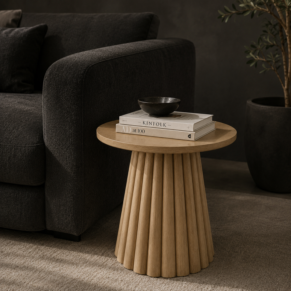 A round light wood side table with a fluted pedestal base, styled in a living room next to a beige sofa with books and a small black bowl on top.