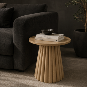 A round wooden end table with a fluted pedestal base, styled in a living room next to a dark gray sofa with books and a bowl on top.