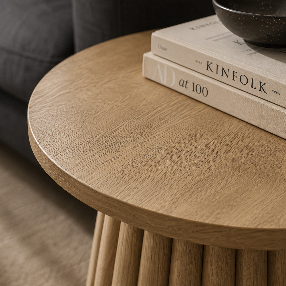A round light wood side table with a fluted pedestal base, styled in a living room next to a beige sofa with books and a small black bowl on top.
